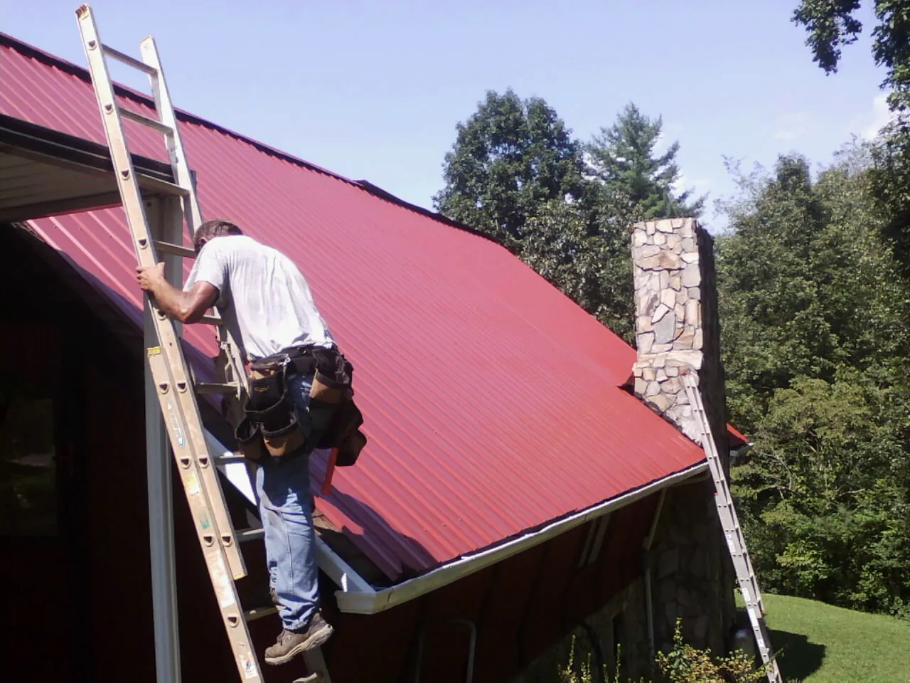 Roofer on a ladder beside a red metal roof and stone chimney — Soffit & Fascia Repair by Roofing Brothers in Beverly Hills